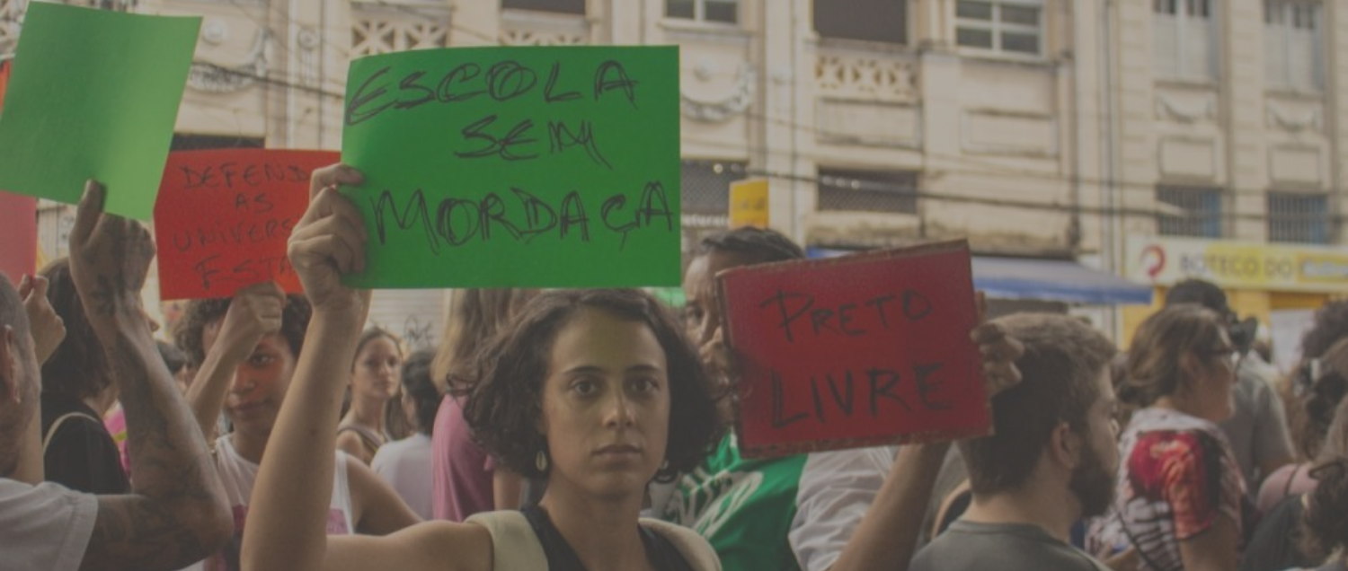 Foto: Midia Ninja || Descrição:Em um protesto, jovens estudantes e professoras erguem cartazes. Em primeiro plano, uma jovem ergue um cartaz verde com o escrito “escola sem mordaça”